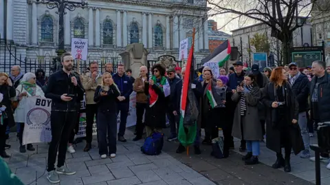 Some of the protesters at the Givan Must Go rally.  A crowd of adults stand in front of Belfast City Hall listening to a man with dark hair and a beard speak into a microphone.  Some of the protesters are carrying Palestinian flags and Givan Must Go placards. 