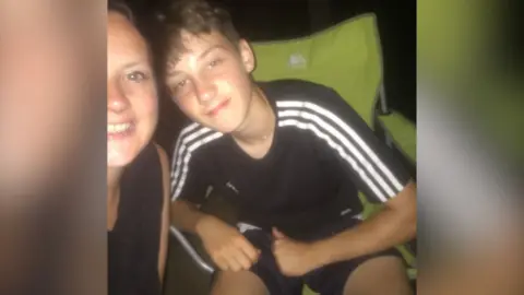 Family handout The photo shows Joshua in a black Addidas t shirt with white stripes on the shoulders. He is sitting in a green camping chair with a small smile on his face, posing for the photo next to his mum.