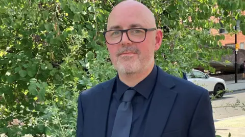 BBC/Victoria Scheer Det Con Ryan wearing a dark suit, dark shirt, and tie stands on a sunlit urban pavement beside leafy greenery, with modern brick buildings, parked cars, and pedestrians visible in the background.