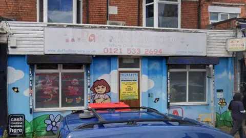 The outside of Daffodils Nursery in Smethwick. The walls are painted in a mural showing blue sky and flowers and a character standing by the door. There is a faded sign above the entrance in Waterloo Road.