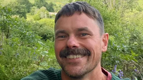 James Dumbelton A man with short brown hair and a moustache smiles at the camera. The background is greenery and foliage.