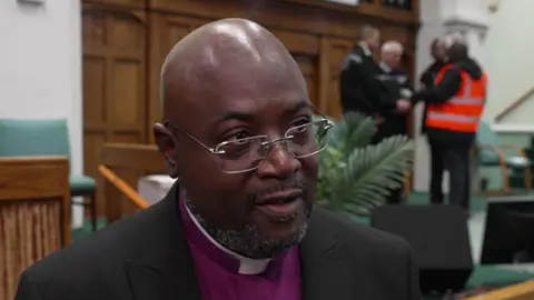 Bishop Tony Parry, wearing a black suit jacket, purple shirt and white clerical collar, speaking in front of a stage with a police officer and a man in a hi-vis jacket on the stage behind him.