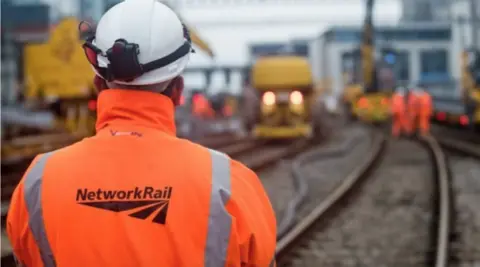 Man with an orange hi-vis Network Rail jacket and helmet stands with his back to the camera looking down a railway line to trains and men a way off