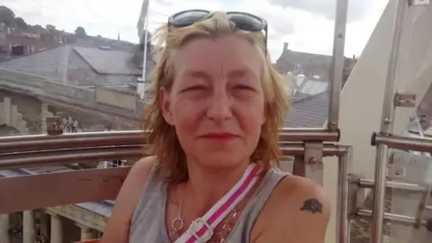 Facebook Dawn Sturgess has short blonde hair and dark sunglasses on her head, She is wearing a grey vest top, silver chain necklace and the pink and white strap of bag is crossing her body. She is sitting on a balcony with rooftops visible in the background qhiqhhidzuidrrprw