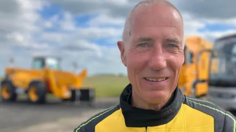 BBC A man looks towards the camera wearing a yellow and black driving suit with the "JCB" logo on the front.