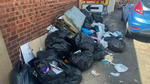 A pile of rubbish on a pavement in Birmingham, with roadwork signs in the background. The rubbish is almost completely blocking the pavement. There is a blue car parked alongside it.