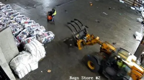 The view from a CCTV camera looking down onto a warehouse floor. There is a yellow machine with prongs on the front, with a person - their face blurred out - standing a distance ahead of it. Large piles of textiles can be seen on the left