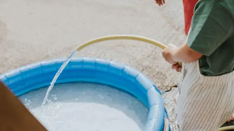 Getty Images Little girl fills a shallow inflatable paddling pool up with water using a yellow hose.
