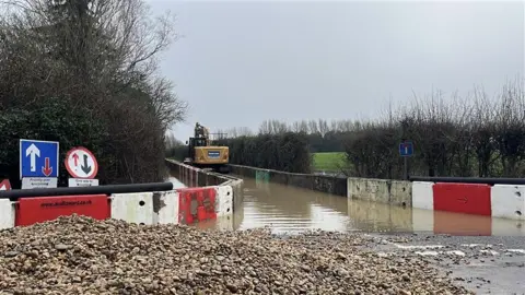 George Carden/BBC A large, yellow digger drives up a flooded road. The road is outlined by white and red temporary fences. There is shingle in the foreground. 