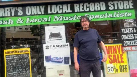 A man in a blue t-shirt, with dark long hair, stands in front of a shop window, with signs show discount record sales amd cpaches to concerts