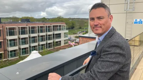 Amy Holmes/BBC Chris Hoggarth standing on a balcony. The balcony is several floors up and overlooks the Red Kite Meadows building, a three-storey development with a row of windows on each floor. Hoggarth, who has short grey hair and is wearing a grey jacket, has his hands on the railing and is turning to look at the camera. 