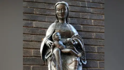 Tiger/Geograph A metal replica of Our Lady of Grace, Nettuno, attached to a brick wall in Ipswich. It shows Madonna wearing a cape and long robe with her feet resting on bricks. On her lap is a reclining infant. 