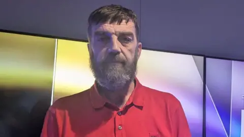Liam is wearing a red polo shirt and standing in a studio with purple lights. He is looking at the camera and has brown hair and a grey beard. 