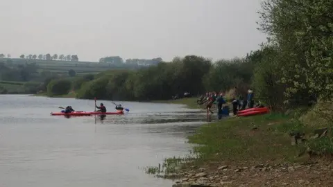 Barry Hurst/Geograph A group of people are in the distance stood on the bank of a reservoir with some canoeing in the river