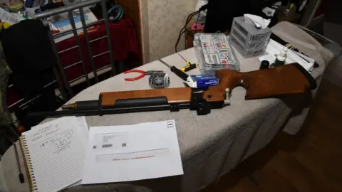 NCA A brown wood and black air rifle lying down on an ironing board which is covered in a grey towel. Next to it is an open notebook with writing in it, and a letter with red text that reads "official notice, investigation opened". there are other items scattered around the gun, including a blue packet or paracetamol, a small pair of pliers with a red handle, a black screwdriver and a plastic container