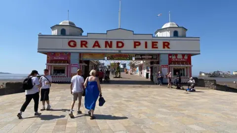 Emma Hallett/BBC People are seen walking into the main entrance of Weston Pier on a sunny day with blue skies overhead.