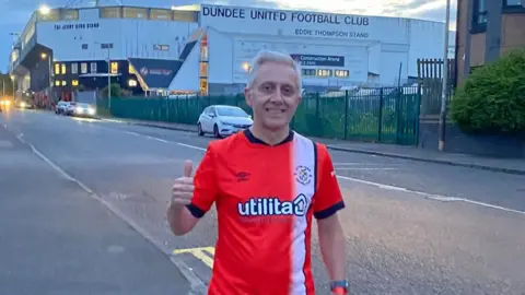 Mark Crowther A man wearing a Luton Town shirt and stood in front of Dundee United Football Club's stadium