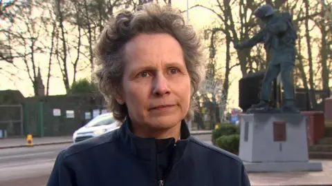 Suzanne Fleming is a woman with short, curly grey hair. She is wearing a navy zip up top which has a Northern Ireland Fire and Rescue Service emblem. She is also wearing a name tag. A statue and a white car are partally visible in the background behind her. 