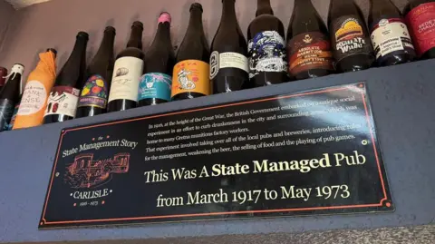 A shelf of a dozen beer bottles, all different brands with different labels. Underneath is a sign stating "This was a state managed pub from March 1917 to May 1973" with information about the state management story in Carlisl