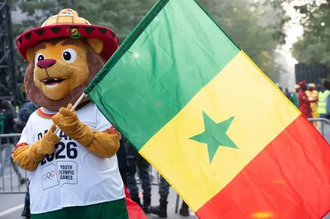 This image shows a friendly-looking lion mascot with a soft, golden-brown face, large expressive eyes, and a wide, cheerful smile. It wears a distinctive, oversized conical hat (a traditional Saloum hat) featuring yellow and orange patterns with a red brim. The mascot is dressed in a white t-shirt that prominently displays the text "DAKAR 2026" above the official Youth Olympic Games logo and holds the national flag of Senegal (red, yellow, and green) in a celebration parade.