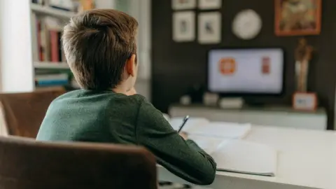 Getty Images The back of a child sitting at a desk looking at a computer screen. Has brown hair, wearing a long sleeve green top and writing in a booklet.