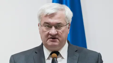 Ukrainian Foreign Minister Andrii Sybiha speaks into a microphone. He has white hair and wears glasses. 