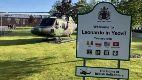 The outside of Leonardo factory in Yeovil. In the foreground, a sign reads "Welcome to Leonardo in Yeovil" and another below reads: "The Home of British Helicopters". In the background, a camo helicopter can be seen on lawn, with the factory behind it.