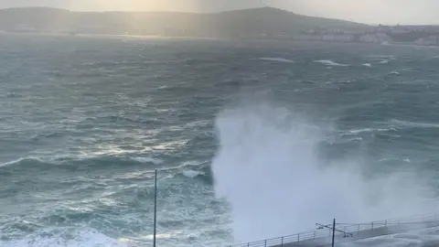 PILGRIMAGE ISLE OF MAN A wave crashes on the northern part of Douglas Promenade, you can see Douglas head in the background, the light is starting to dwindle.