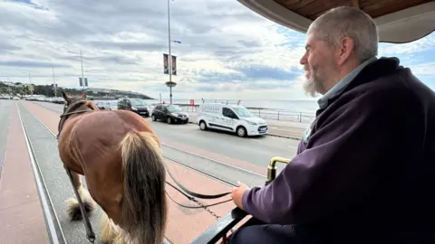 Peter Cannon on the left is at the front of the tram holding the reins of the brown horse on the left walking along Douglas Promenade with cars in the background.