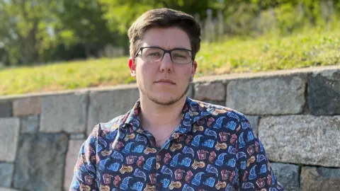 Sam is wearing glasses and a patterned button-up shirt sits on a wooden bench beside a stone wall in a sunny park, with green grass and trees in the background.