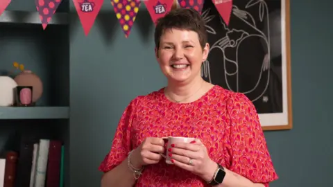 Supplied A woman with short brown hair wearing a pink and red dress holds a pink mug. She is standing in a living room with dark teal walls and above her is pink bunting.