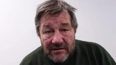 A man of around 60 pictured in a police mugshot. He appears to be hunched, has grey hair and a short beard and is wearing a dark top.