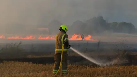 A firefighter with his back to the camera trains a hose on a smouldering field. In the distance, beyond him, another firefighter is tackling a line of flames. The air is thick with smoke.