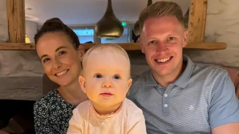 Harry Foster is pictured wearing a blue polo shirt and smiling at the camera. Sat beside him is his wife, who has long brown hair and a black and white patterned shirt and in front of them is their baby who has fair hair.