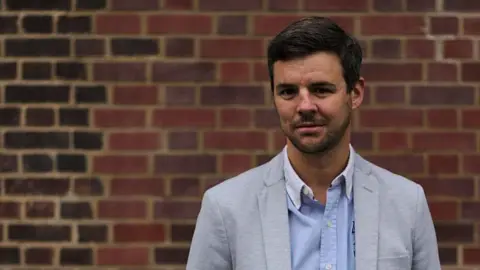 Seb Pelisse Seb Pelisse, who has dark, short hair and facial hair, stands in front of a brick wall smiling. He is wearing a blue shirt and light grey jacket.