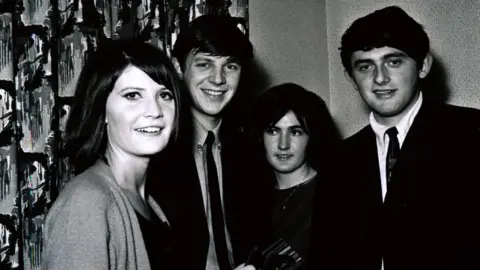 Gliderdrome Black and white photo of sanide shaw signing autographs. in the background are two men in 60s style suits and a woman wearing a dark dress