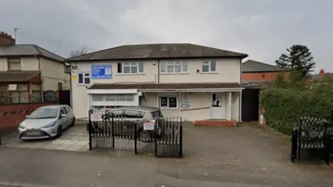 General view of the entrance of Naseby Medical Centre in Birmingham. A cream coloured building with a small driveway outside. Two cars are parked in the driveway which has some iron gates across part of it.