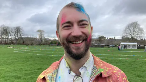 Will Stone smiles broadly. He is wearing an orange jacket and white shirt and is covered in brightly coloured paint powder.