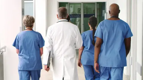 Three health care staff with their backs turned to the camera walk down a long corridor wearing blue scrubs. A fourth worker stands in the middle with his back to the camera wearing a long white coat and holding a clipboard.