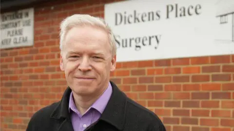 Jamie Niblock/BBC A man is wearing a black jacket over a purple shirt. He has short grey hair and is standing in front of a brick GP surgery.