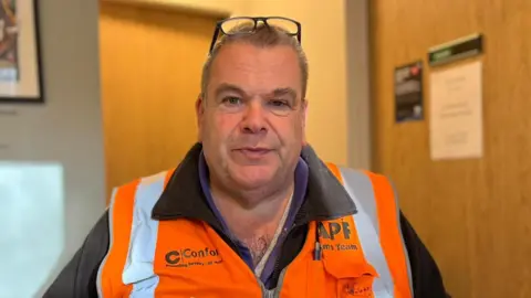 Lachlan McEwan, in a black jacket and orange hi-viz vest and glasses on his head, looks at the camera. He appears to be in an office and there is a door behind him with notices on it.