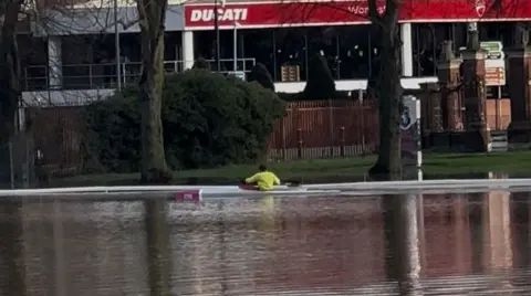 Seb Sargent/BBC A rower, pictured from the back, rowing on water, with a garage, trees and railings pictured in the background.
