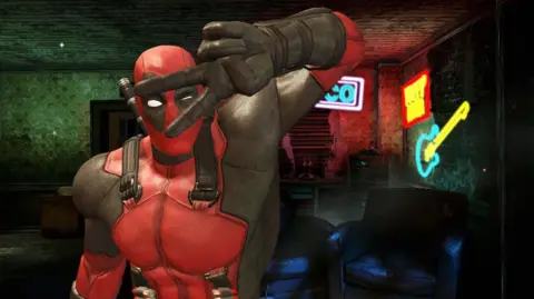 Activision/Marvel A screenshot of Deadpool from the 2013 computer game showing the peace sign with his fingers.  He is a superhero dressed in a red suit with black over the eyes and gloves.