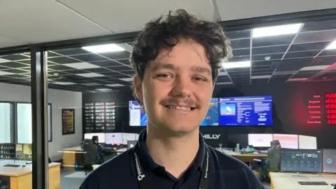 A picture of Oliver smiling in a room. He is wearing a black shirt and has black hair.