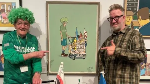  Fundraiser John Burkhill is standing on the left of the picture with his pram in front of him pointing to a painting of him which is being raffled off. On the right is the Sheffield artist Pete McKee, he is wearing brown trousers, and a dark-green checked shirt. He has short light-brown hair and is wearing dark-rimmed glasses. 