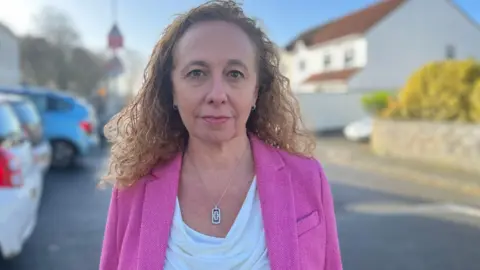 Inna Gardiner is standing in a street with houses behind her. She is wearing a pink jacket and has shoulder length brown curly hair.