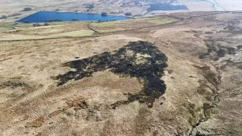 Overhead shot over Ralfland Common, near Shap, showing a large, blackened area where the fire damaged grasslands. The pictured landscape is predominantly brown, with some treees surrounding a lake in the distance and a small river running to the right of the image.