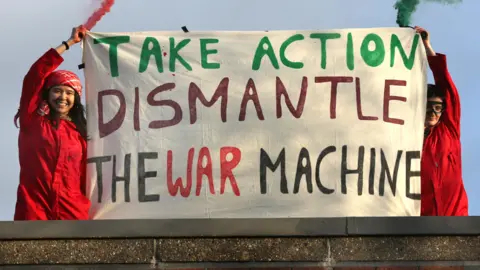Audrey Corno and Jessica Stacey, holding up their arms and smiling, wearing red jumpsuits. The woman on the left has dark hair and has a red and white scarf tied around her head. The woman on the right has black goggles on. There is a banner in the middle of them that reads "Take Action Dismantle The War Machine".