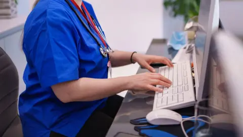 An anonymous nurse wearing blue scrubs and with a lanyard and stethoscope around her neck sits at a computer at a desk in what appears to be a medical room.