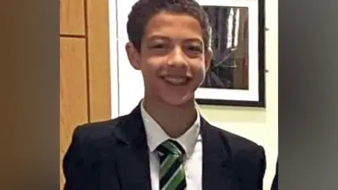 Noah Donohoe, a boy with dark hair, wearing a black blazer, white shirt and green, black and white tie.